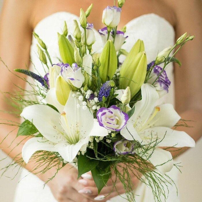 Bathed in the gentle light of a Brent Cross morning, this bridal bouquet stands as a radiant testament to local floral artistry. At its centre, robust white Oriental lilies open to reveal dustings of petite speckles and sunlit yellow-orange stamens. Interspersed gracefully among them, lisianthus blooms display their crinkled, ruffled textures, their edges tinged in a soft lavender that recalls the fleeting pastel hues found in Brent Reservoir at dusk. Arresting accents of deep violet Veronica lend the bouquet a modern edge, while miniature white baby's breath offers airiness, resembling delicate clouds drifting across the composition. Wisps of asparagus fern and broad, deep green leaves provide layers of freshness, spilling gently beyond the frame and echoing the natural lines of local North West London parks. Gently manicured hands, brushed in soft neutral polish, hold these artfully wrapped stems, grounding the bouquet in intimate anticipation. The soft-focus folds of the bride's strapless gown create a tranquil canvas, allowing the floral arrangement to shine brightly against its serene backdrop. Crafted for wedding ceremonies near Brent Cross Shopping Centre or tucked-away synagogues, this bouquet celebrates both love's tradition and individual expression, blending romance, elegance, and a flourish of local character.