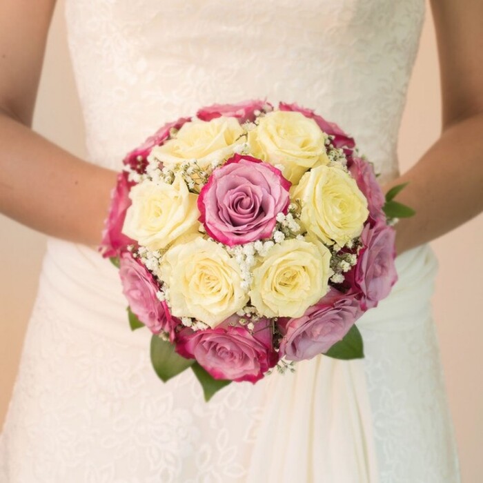 This evocative bridal bouquet creates a stunning focal point, lovingly hand-tied by a Brent Cross florist deeply rooted in the traditions of North West London. A mix of full, creamy yellow roses and bold, fuchsia pink blooms form the central structure, with touches of mauve catching the light in subtle gradations. Wisps of gypsophila thread through the bouquet, their star-like blossoms lending delicacy and volume. Rich green leaves peek from the boundaries, framing the flowers. The bouquet is cradled by the bride at waist height-her arms relaxed, her wedding dress an ivory canvas showcasing both intricate lace across the bodice and a newly draped, flowing skirt. Sunlight enhances the detail in both petals and fabric, casting gentle shadows and revealing the craftsmanship in every element. Separated from the world by a blurred, pale background, the arrangement radiates joy and meticulous artistry. It conjures images of celebrations near Brent Cross Shopping Centre, garden parties, or intimate ceremonies along Hendon Way-occasions where every petal is chosen to evoke romance, anticipation, and a quiet sense of belonging.