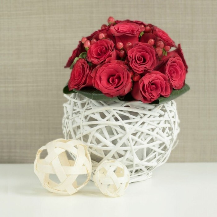 An artfully composed dome of crimson roses sits boldly atop a sculptural white wicker sphere, evoking the feel of an intimate wedding table setting in Brent Cross. The roses create a compact, cushion-like bouquet, each head nestled closely to the next, forming a rich tapestry of velvety petals. Colour ranges from deep wine-red in the shadowed centres to brighter, glowing crimson on the outer petals where the light touches. Some roses remain shyly furled, while others open broadly, their layered petals spiralling outwards in soft waves. Threaded between these blooms are glossy red hypericum berries, smaller and rounder than the rose heads, their surfaces catching the light in tiny highlights that add vibrancy and dimension. A neat band of dark green foliage forms a discreet collar at the base, just visible beneath the flowers and giving a gentle, natural contrast. Supporting the arrangement is a distinctive spherical vase made from thin white-painted twigs, woven and interlaced into an open globe. The gaps in the weaving allow the surrounding light to enter, creating delicate patterns of shadow on the gleaming white tabletop below. To the left of the main arrangement, two decorative wicker balls in a soft, natural beige stand as playful echoes of the vase, one larger sphere placed slightly in front of a smaller one. Their casually tangled strips of wood bring an easy, organic feel, reminiscent of relaxed interiors in modern flats near Brent Cross Shopping Centre or along Hendon Way. Behind the display, the background dissolves into a softly blurred canvas of pale taupe or linen beige, offering just enough texture to be interesting without drawing the eye from the flowers. The overall impression is of refined romance and contemporary style, making this design perfectly suited as a wedding table arrangement, anniversary centrepiece, or special occasion display prepared by Brent Cross Flowers for local celebrations.