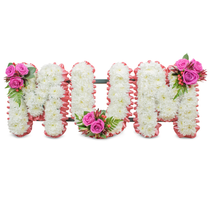 A poignant, handcrafted memorial piece spelling out MUM in bold capital letters, each letter densely composed of small, immaculate white chrysanthemums that form a plush, even surface. The arrangement sits against a plain white field, allowing every textured petal and layered bloom to read clearly. Around the perimeter of each letter a softly ruffled pale pink fabric or ribbon traces the shape, adding a touch of maternal warmth and a subtle colour halo that lifts the otherwise pure white palette. Structural support appears as a dark green frame between the letters, discreet yet visible, holding the tribute upright for display at a service or graveside. Three carefully positioned clusters of accent flowers punctuate the design: the first M is accented at its upper left by several deep fuchsia roses in various stages from bud to open bloom, interlaced with glossy green fern-like leaves and small bright hypericum berries; the U bears a matching grouping at the base of its curve, and the second M mirrors the first with another bouquet on its upper right. The combination of crisp chrysanthemums, the velvety texture of roses, and the subtle sheen of berries creates varied tactile impressions; light catches the rose petals and the ruffled ribbon differently, giving a soft relief across the surface. The overall mood is one of reverent affection - a traditional funeral tribute chosen to honour a mother with dignity and love. As a local florist-crafted piece suitable for services near Brent Cross Shopping Centre, Hampstead Garden Suburb or Cricklewood, it conveys comfort through classic sympathy symbolism and considered floral workmanship.