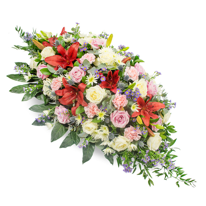 A large, meticulously arranged funeral tribute is set against a plain white background, its elongated shape and generous mix of blooms evoking both tradition and gentle modernity. The spray has a graceful, tapering form, fuller through the centre and narrowing at each end, so it could comfortably sit along the length of a casket during a service in Brent Cross. At its heart, deep red lilies with wide, star-like petals open outward, their rich tone and speckled throats giving the arrangement a strong focal point and a sense of gravity. Surrounding these are clusters of roses in creamy white, soft blush, and pale pink, each bloom at a slightly different stage, from bud to full blossom, adding depth and a natural, unforced rhythm to the piece. Light pink carnations, with their soft, ruffled edges, nestle between the larger flowers, while delicate white spray chrysanthemums provide clusters of small, many-petalled blooms that fill in the spaces and lend a feeling of completeness. Interspersed throughout the spray are muted lavender and purple filler flowers, likely statice or veronica, as well as a few striking blue thistle-like stems, whose spiky, architectural form and cool hue introduce a refined contrast. Beneath it all lies a rich bed of greenery composed of broad, polished leaves and finer, airy foliage that extends slightly past the mass of flowers, softening the outline and giving the spray a natural, flowing silhouette. The overall impression is one of serene remembrance and steady comfort, the type of thoughtful floral arrangement a family might receive from Brent Cross Flowers when saying goodbye to a loved one at a nearby crematorium or place of worship.