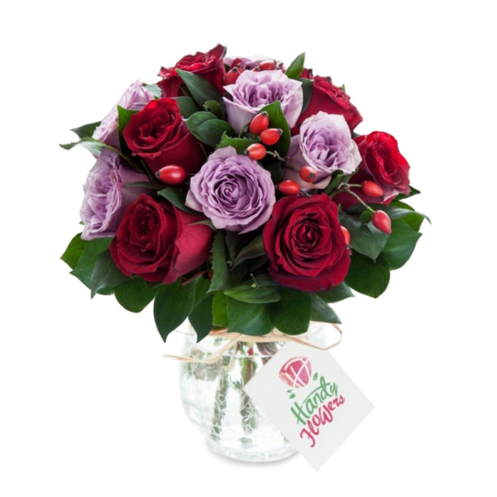 A thoughtfully composed, round arrangement of roses in saturated colour: deep crimson and burgundy roses sit alongside delicate lilac and dusty purple varieties, all captured mid-bloom with petals gently unfurling. The grouping is dense and sculpted, the eye guided by the alternating dark reds and soft purples, while robust dark green foliage - likely Ruscus or Salal - fills the gaps and provides structural contrast. Bright orange-red rose hips are artfully nested throughout, offering small pops of colour and a rustic texture that plays off the refined velvet of the roses. The bouquet is presented in a clear, antique-style mercury glass vase that is bulbous at the base and narrows toward the top; its silvered, crackled finish scatters highlights and lends an old-world shimmer. Around the neck a short length of natural twine or raffia is tied, from which hangs a small rectangular white tag printed with a simple rose motif and room for a personal message. Photographed on a stark white background, the arrangement reads as premium and freshly gathered, the colours crisp and true. As a local florist I picture this piece being offered for delivery by Brent Cross Flowers to homes across Brent Cross, Cricklewood and nearby Golders Green - ideal for anniversaries, meaningful thank-yous, or as a centrepiece that lifts a quiet breakfast table with scent and colour.