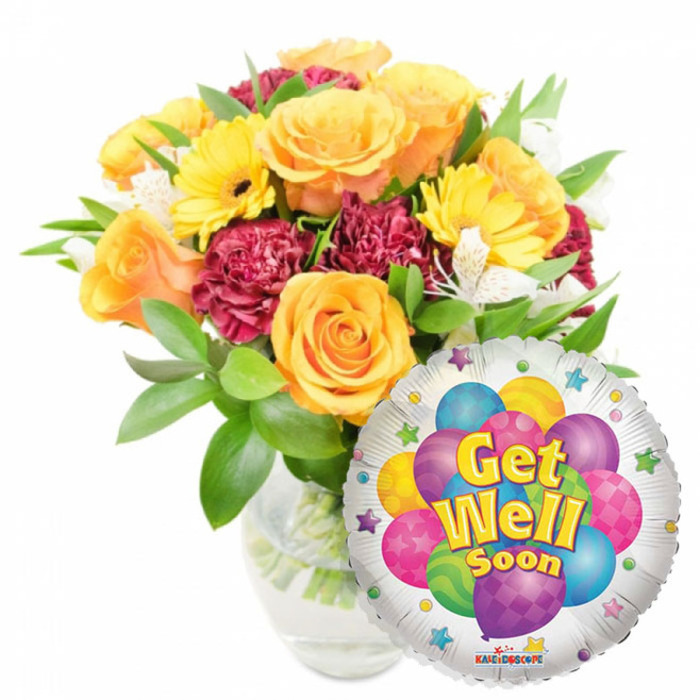 A heartfelt, uplifting arrangement designed to comfort and encourage: a full bouquet of fresh flowers sits in a clear glass vase and is paired with a round metallic balloon bearing a heartfelt message. The floral palette is warm-sunny yellows, soft oranges and deeper red-burgundy tones combine to create a lively yet soothing effect. Several opening orange and deep yellow roses dominate the bouquet, their spiralling petals giving a sense of classical beauty and gentle motion. Bright yellow gerbera daisies provide bold faces and clean lines, while ruffled maroon or burgundy carnations add density and textured shadow between the larger blooms. Small white alstroemeria (Peruvian lilies) are tucked through the mass of colour, offering fine, delicate accents that lighten the visual weight. Lush green foliage frames and supports each blossom, its varied leaf shapes adding depth and a fresh, living backdrop. The glass vase is clear, the stems and water visible beneath, lending an honest, simple presentation that keeps the focus on the blooms. In front of the lower right of the bouquet a shiny silver Mylar balloon partially overlaps the flowers; its centre shows a lively cluster of cartoon balloons in bright purple, sky blue, hot pink, yellow and lime green, some patterned with stripes or dots, while bold yellow-and-blue letters outlined in white read Get Well Soon. Tiny starburst motifs scatter around the balloon's white border and a small KALEIDOSCOPE logo is visible at the bottom edge. Bathed in soft natural light, this composition would lift spirits for a loved one recovering in Brent Cross, the surrounding Cricklewood and Hendon neighbourhoods, ideal for hospital or home visits.