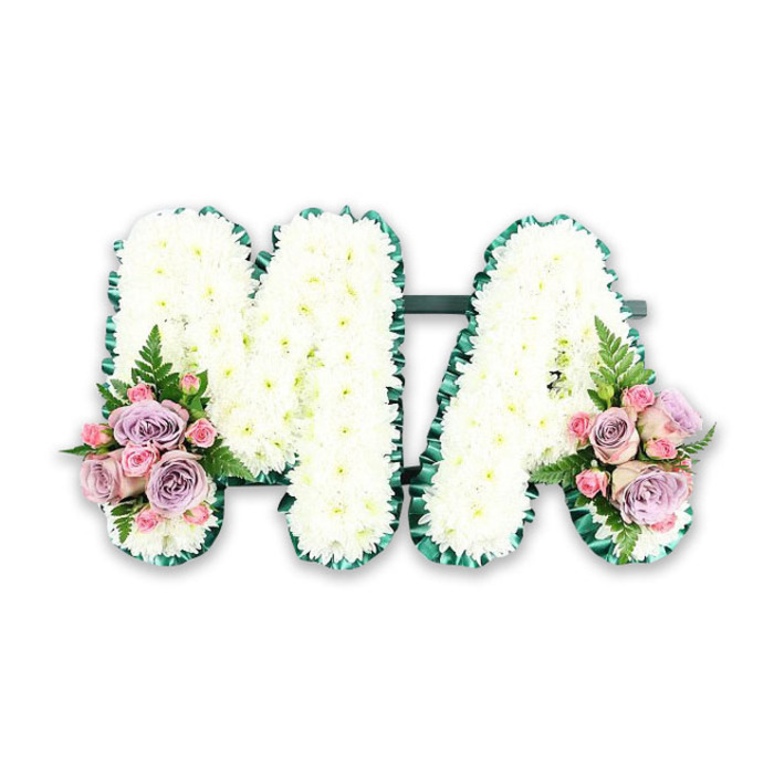 A meticulously arranged funeral letter tribute spells out MA in upright capitals, a classic expression of love for a mother or maternal figure. The structure is entirely concealed beneath a thick covering of white chrysanthemums, their small, cushion-shaped blooms pressed close together so that only the soft texture of petals can be seen. The flowers create an almost quilted effect across the surface of each letter, giving the design a sense of comfort and serenity. Tracing the outer edges of the M and the A is a lush band of deep green satin ribbon, pleated in fine ruffles. This decorative border adds a subtle sheen and carefully sharpens the silhouette of the letters, making them clearly legible even from a short distance. The two letters are linked by a slim, darker green crossbar, discreetly placed so the composition feels solid and unified. At the base of the left leg of the M and the right leg of the A, dainty accent sprays lend colour and emotion. Each cluster blends soft lilac roses with blush and baby-pink roses, including smaller spray roses that add variety in size and form. Their petals, satiny and gently curved, contrast beautifully with the crisp chrysanthemums. Feathery fern fronds and sprigs of fresh greenery peek out at the edges, bringing lightness and movement to the piece. Photographed against a stark white background, the tribute appears calm and composed, making it suitable for a funeral or memorial in Brent Cross, Cricklewood, or along the Edgware Road. The overall impression is of quiet grace and heartfelt remembrance, a floral message that simply spells Ma with love.