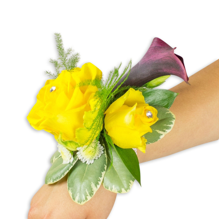 Captured on a pale to medium forearm against a stark white background, this wrist corsage combines sunny warmth and moody depth in a compact, wearable design. The centrepiece comprises two vibrant yellow roses - the left bloom opened wide, petals gently rolling back, and the right blossom more cupped and intimate - each accented with a tiny clear rhinestone placed near the centre to introduce a refined sparkle that catches the light. Prominent behind the roses and oriented slightly upward is a single dark plum calla lily; its smooth, trumpet-like silhouette and deep, near-black inner tones create a strong visual anchor and an elegant counterpoint to the roses' cheerful hue. The floral trio is nestled within a variety of green textures: delicate asparagus fern lends a soft, airy veil around the top and background, while larger variegated leaves with pale margins (reminiscent of pittosporum) form a stable, glossy base. Small clusters of white filler flowers - petite chrysanthemums or tiny spray blooms - fill gaps and impart a meadow-like fullness. A thin, concealed band secures the corsage to the wrist so the flowers appear naturally placed rather than floating. Lighting is neutral and bright, chosen to reveal the tactile contrast between the rose petals' velvety layers, the calla's satiny surface, and the foliage's mixed textures. The effect is wearable elegance suitable for proms, weddings, or celebratory evenings across Brent Cross, from a rehearsal dinner near the shopping centre to a wedding photo by nearby Silver Jubilee Park, offering a joyful yet sophisticated accent to a formal outfit.