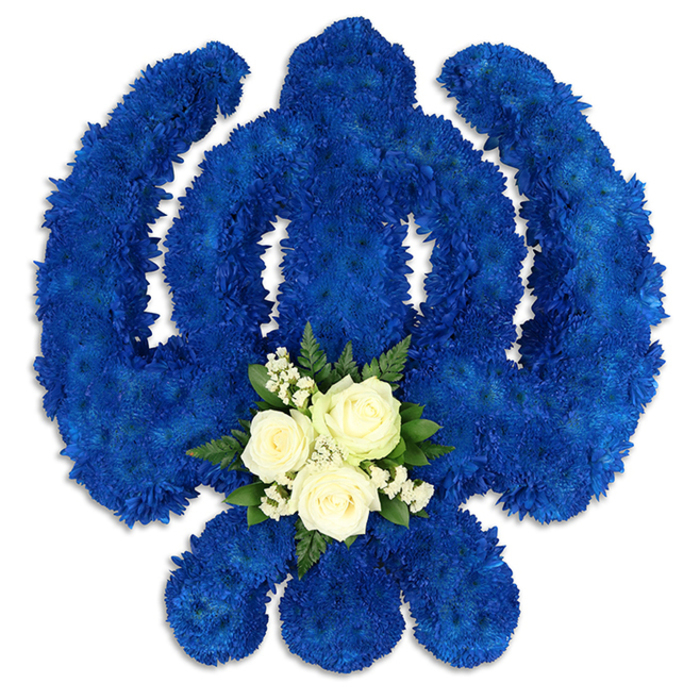 Presented against an immaculate white backdrop, this large Khanda floral tribute appears both commanding and tender, an unmistakable symbol of Sikh faith interpreted in flowers. The entire emblem-the tall central sword, the circular chakkar that surrounds it, and the two protective kirpans that curve at its sides-is painstakingly formed from countless royal blue chrysanthemum florets. Each small bloom is nestled closely to the next, creating a dense, velvety field of blue with a uniform depth of colour. Soft light glances across the petals, revealing fine textures and giving the structure a sculpted, three-dimensional feel. At the base of the central circular section, a serene accent of white and green provides balance and focus. Here, three white roses sit together, their petals delicately layered and just beginning to unfurl, suggesting gentleness and heartfelt remembrance. They are encircled by sprigs of tiny white gypsophila, whose airy flowers lighten the composition, while deep green foliage creates a natural frame and introduces a soothing, earthy tone. This central cluster seems to rest like a quiet offering placed within the heart of the Khanda. Subtle shadows beneath the design indicate that it has height and presence, as though arranged on an easel or stand for viewing during a service. Crafted with the experienced hand of a florist serving Brent Cross and surrounding neighbourhoods along the North Circular and near Brent Cross Shopping Centre, this floral Khanda is a thoughtful choice for Sikh funerals, memorials, and commemorations. It conveys respect, devotion, and the enduring comfort of tradition.