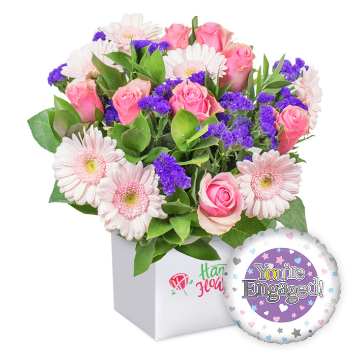 A sparkling engagement bouquet sits in a clean, white square box, ready to celebrate a couple's special milestone in the Brent Cross area and beyond. The arrangement bursts with several full-bloom pink roses, their petals opening in classic romance, nestled among cheerful light pink and white gerbera daisies that paint the display with their generous, sunlit petals. Delicate clusters of vivid purple filler flowers peek between blooms, adding depth and a whisper of whimsy, while abundant green foliage provides a lush, natural backdrop that enhances the bouquet's freshness. The container is a modern, white square box with a gentle, understated logo on the front, featuring a red rose symbol accompanied by green script-an elegant hallmark of quality and local craft. To the lower right, a shiny foil balloon completes the set, its design centered on a purple circle bearing the words You're Engaged! in refined silver lettering, surrounded by a white ring decorated with tiny pastel hearts and silver stars. Perfect for congratulating a fiancée or newly engaged couple, this gift exudes joy, romance, and warmth, and is well-suited for delivery across Brent Cross, Cricklewood, Golders Green, and Hendon, bringing a touch of ceremonious charm to any home.