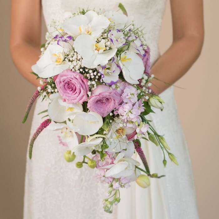 A bride from Brent Cross stands serenely, the photograph capturing her from mid-torso as she holds an extraordinary cascading bouquet-a true celebration of both artistry and locale. The bouquet commands attention: white Phalaenopsis orchids, iconic for their pure elegance, flow downward in gentle arcs, their centers tinged with gold. Around them, roses in shades of pale blush and lavender create a poetic interplay of colour and texture, each petal touched by natural light reminiscent of morning in Princes Park. Baby's breath and interspersed buds give a light, airy effect, softening the bouquet's silhouette. Textural drama arrives through bold, magenta amaranthus, trailing in soft, feathery tendrils that contrast with slender green foliage and unopened buds tucked within. The bride's fingers delicately cradle this lush creation over a gown detailed in fine lace, her presence bringing intimacy and meaning to the scene. The neutral background, suggestive of sunlit interiors found in Brent Cross homes, lets the florals and craftsmanship stand in tranquil relief. This bouquet, brought to life by Brent Cross Flowers, embodies the hope and excitement of wedding celebrations here in North London, offering inspiration rooted in both elegance and local flair.