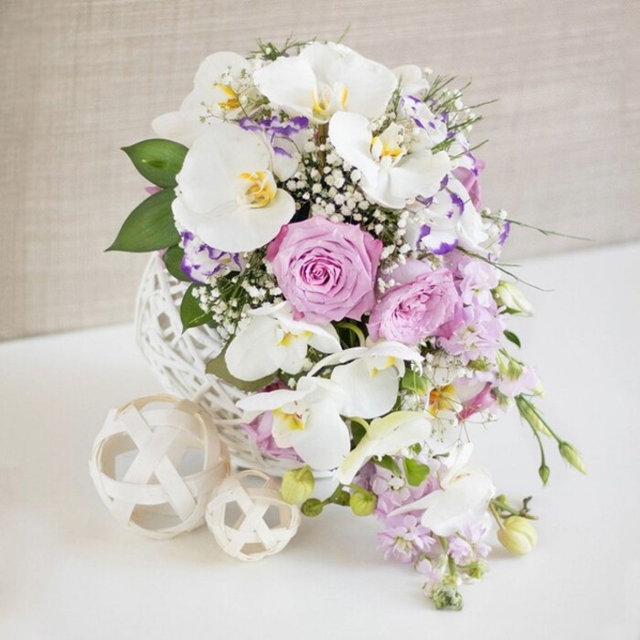 An image of quiet sophistication shows a meticulously arranged floral centerpiece placed on a smooth white table, reminiscent of a wedding reception near Brent Cross or a refined celebration in nearby Temple Fortune. The arrangement is rooted in a distinctive, spherical vase constructed from white woven strands that crisscross to form an open, airy lattice, much like an oversized decorative wicker ball. From this sculptural base, a lush bouquet rises and gently spills outward in a rounded silhouette. Dominating the display are several large white Phalaenopsis orchids, their broad petals smooth and slightly curved, each bloom featuring a yellow and golden centre that adds depth and a hint of warmth. Tucked around these orchids, generous roses in pastel pink and soft lavender tones blossom fully, their ruffled petals layered densely to create an impression of richness and romance, ideal for a bridal table close to Brent Cross station. Intertwined with the larger flowers, sprays of white baby's breath scatter like tiny stars, knitting the arrangement together and adding a light, cloud-like softness. Additional smaller blossoms edged in purple and fuchsia-resembling Lisianthus or Eustoma-bring subtle colour variation and fine, ruffled texture. A few slender stems bearing pale lilac blooms trail down from the bouquet, providing gentle movement and a hint of cascading elegance. Throughout the design, polished green leaves and more delicate, fern-like foliage add contrast and shape, ensuring the pastel shades remain vibrant rather than pale. Resting nearby on the same white surface, two decorative woven balls in a cream or natural tone-one slightly larger, one smaller-echo the texture and form of the vase, creating a cohesive, contemporary look. Against a softly textured neutral beige background, slightly out of focus, the entire composition feels calm, bright, and celebratory, perfectly suited to an elegant Brent Cross wedding table or special occasion setting.