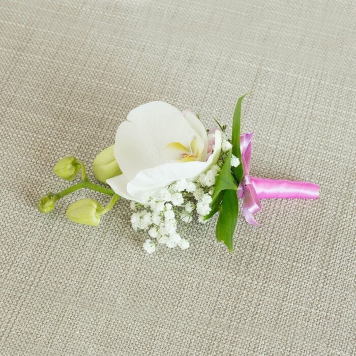 A finely crafted floral boutonniere lies horizontally across a light oatmeal-toned linen fabric, the natural weave of the cloth visible as a gentle, textured backdrop. At the centre of this small composition is a single, immaculate white Phalaenopsis orchid, its petals broad, smooth, and slightly arched, like porcelain wings unfolding. The orchid's throat reveals delicate details: a soft wash of yellow, tiny touches of pale pink, and subtle markings that draw attention inward, creating a refined focal point. Surrounding the base of the orchid, a mass of tiny white baby's breath blossoms forms a cushion of airy, cloud-like texture, their countless miniature blooms lending an almost misty softness to the arrangement. A few slender, bright green leaves emerge from behind the flowers, providing a fresh contrast that keeps the boutonniere from feeling too monochrome. Off to one side, a short branch bears a couple of small, tightly closed lime-green orchid buds, their smooth surfaces and rounded shapes suggesting the quiet anticipation of future blooms. The stems of the flowers and foliage are gathered neatly and wrapped in a lustrous fuchsia pink satin ribbon, tied into a compact, elegant bow with short, tidy tails; the ribbon's vivid hue and glossy finish introduce a joyful burst of colour against the neutral fabric and white petals. The lighting is soft and diffused, enhancing the velvety feel of the orchid, the fine granularity of the baby's breath, and the tactile quality of the linen. This boutonniere exudes classic, understated elegance, an ideal finishing touch for a groom or groomsman at a Brent Cross wedding, a school prom nearby, or a formal celebration hosted in one of the local hotels or event spaces that line the surrounding North London streets.