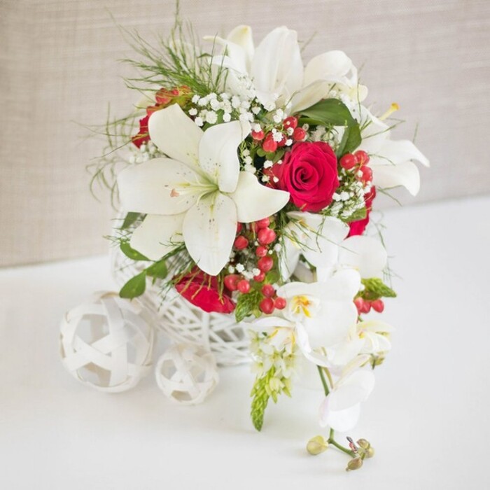 An elegant flower arrangement in a refined palette of white, crimson, and fresh green is displayed on a white tabletop, as if styled for a special celebration overlooking Brent Cross. The centerpiece is contained in a round, open-lattice vessel crafted from white, woven material that resembles fine wicker or rattan, giving the design a light, modern feel. Emerging prominently from this basket are several large white oriental lilies, their petals broad, slightly curved, and silky in texture, with prominent stamens dusted in warm golden pollen that hints at their softly perfumed presence. Cradled among the lilies are rich red roses in various stages of opening, from tightly furled buds to partially open blooms, their velvety petals adding depth and romance. Delicate sprays of white baby's breath are threaded throughout, creating a misty, cloud-like effect that gently unites the different flowers. Wisps of fresh green foliage, fine and feathery, peek out between the blossoms, lightening the composition and suggesting a freshly arranged piece straight from a local Brent Cross Flowers workroom. Punctuating the design, clusters of bright red hypericum berries add glossy, rounded accents that feel festive and lively. Extending gracefully from one side of the arrangement, a cascading stem of white Phalaenopsis orchids flows outward, each bloom cool and pristine with a hint of yellow and blush-pink at the center, followed by a line of small buds. To the left of the main bouquet, three decorative white woven spheres sit on the tabletop, echoing the container's lacy structure and balancing the visual scene. The background is a soft, light beige fabric with subtle texture, ensuring that the flowers remain the focal point while evoking a calm, luxurious atmosphere ideal for wedding tables, anniversaries, or elegant gatherings in Brent Cross.