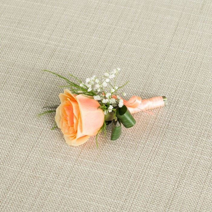 A delicately styled peach rose boutonniere rests lengthways on a soft, light beige fabric with a distinct linen-like weave, the neutral background giving the flowers a calm, timeless setting. At the centre of the composition is a single, partially opened rose, its petals forming a graceful swirl that deepens from a warm, apricot-peach at the heart to a pale, creamy blush at the outer edges. The rose appears freshly cut, with petals that look velvety and cool to the touch, suggesting a light, sweet fragrance. Nestled to the upper right of the bloom is a small cluster of white baby's breath, each minuscule blossom dotting the arrangement with airy flecks of white, adding both texture and a romantic, bridal quality. Slender, grass-like green sprigs and a couple of broader, darker green leaves extend from behind the rose, the larger leaves gently curved to cup the stems and lend structure. The stems themselves are carefully bound in a peach satin ribbon that mirrors the tones of the flower, the ribbon wrapped smoothly and finished in a discreet bow or knot at the base, showing the precise hand of an experienced florist at Brent Cross Flowers. Soft, even lighting washes over the scene, picking up the satin sheen of the ribbon, the subtle shadows in the rose's layered petals, and the fine grain of the woven fabric below. The overall impression is of refined, effortless elegance, perfect for a groom or guest at a wedding near Brent Cross Shopping Centre, a registry office ceremony, or a formal event hosted in one of the nearby North London venues, where a small yet meaningful floral detail completes the look.