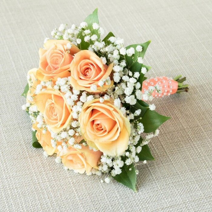 Laid horizontally on a light beige, linen-like background often found in welcoming Brent Cross homes, this exquisitely curated bouquet conjures thoughts of bridal parties mingling along bustling High Road or in sunlit local gardens. At its heart, seven to nine generously bloomed roses bloom in seamless peach and apricot gradients, their softly layered petals opening outward with innate grace. Dispersed artfully among the showstopping roses are delicate tufts of white baby's breath (Gypsophila), infusing the composition with an airy, ethereal quality akin to wisps of early morning fog drifting past Brent Park. Punctuating the arrangement are deep green rose leaves, their glossy surfaces lending structure and the freshness of a local florist's early-morning deliveries. The bouquet's rounded silhouette is achieved through careful artistry, its stems wrapped tightly in a coral-peach satin ribbon that's both tactile and visually harmonious with the flowers above. Further elevating the handle's beauty, a line of small, pearlescent white faux pearls is meticulously coiled around the ribbon, imparting a sense of vintage glamour and a subtle sparkle under soft lighting. The neatly trimmed stems are just visible at the ribbon's end, emphasizing the bouquet's hand-crafted authenticity. With each detail bathed in gentle, even illumination, this arrangement is ideally suited for a chic Brent Cross wedding, a memorable engagement celebration, or as an elegant gift echoing the heartfelt traditions of the local community.