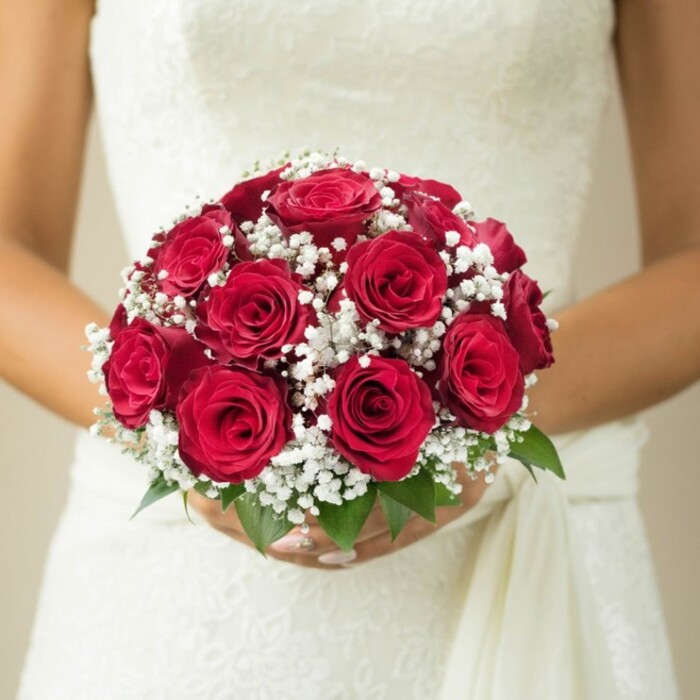 Framed against a softly blurred cream backdrop reminiscent of Brent Cross's stylish interiors, this close, mid-torso image captures a bride immersed in a moment of grace. The focal point is a standout bridal bouquet: an opulent dome of deep, burgundy red roses, their velvety petals expertly arranged to showcase every stage of blossoming. Dotted generously with clouds of pure white gypsophila, the bouquet radiates a light, ethereal contrast, making the reds glow with intensity. Sprigs of fresh green foliage at the base evoke the nearby parks and gardens that define much of Brent Cross's neighbourhood charm. The bride's gentle, manicured hands embrace the bouquet, signifying both personal care and the skilled touch of a local artisan florist. Her wedding gown, glimpsed behind the arrangement, seems crafted with textured lacework along the bodice and a flowing sash that adds a note of understated luxury-echoing the quiet elegance found in Brent Cross's community gatherings. The scene is ripe with anticipation-the essence of wedding celebration distilled into a single floral moment, ideal for guiding couples as they select their bridal blooms amid the welcoming streets of North West London.
