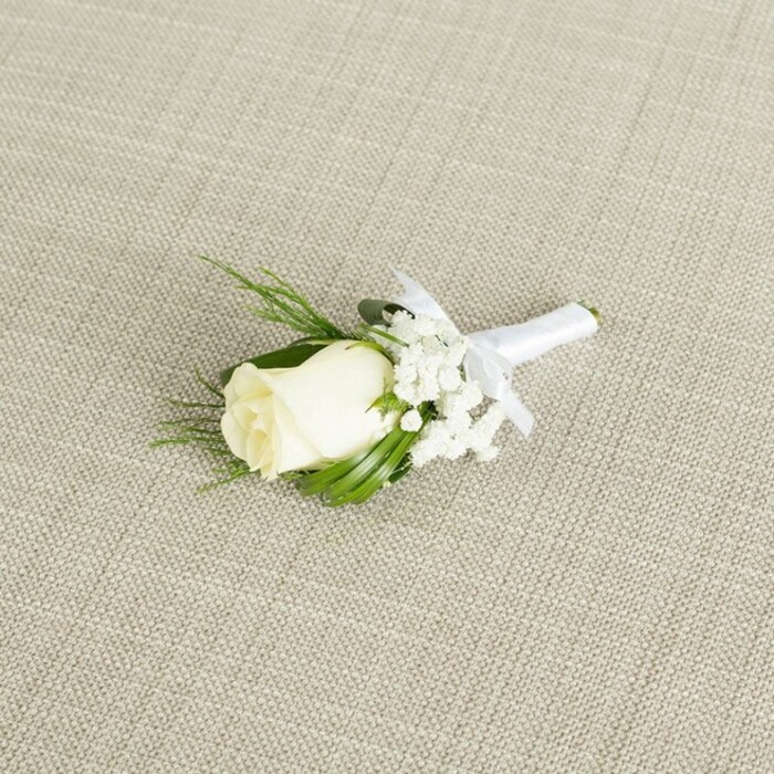 A refined white rose boutonniere is laid diagonally across a softly textured, light beige background, reminiscent of natural linen or finely woven cotton often seen on elegantly set tables at Brent Cross wedding receptions. The central focus is a single creamy white rosebud, its petals tightly yet gently furled, suggesting freshness and a sense of poised anticipation, like a moment just before the vows are spoken. The rose's velvety petals reveal a subtle gradation from warm ivory at the core to a softer, milky white at the edges, each fold catching the gentle, diffused light. Nestled to one side and partially behind the bloom, airy clusters of white baby's breath offer a delicate, cloud-like counterpoint, their tiny, rounded flowers creating a soft halo that enhances the rose without overpowering it. Slim, dark green leaves and a few fine, feathery sprigs of foliage cradle the composition, their varied textures-from glossy to slightly matte-adding depth and a natural, garden-inspired feel that might evoke quiet corners of Clitterhouse Playing Fields or tree-lined streets near Brent Cross. The stems of the rose and greenery are neatly gathered and wrapped with pure white satin ribbon, tightly and evenly wound, with a small, graceful bow tied just beneath the flower head. The bow's loops are carefully shaped, giving the impression of an experienced florist's hand and thoughtful preparation for a lapel, whether for a groom, best man, or proud father. The background fabric's diagonal weave is clearly visible, providing an understated, tactile context that makes the whites appear even more luminous. Soft, even lighting enhances every detail-the chiseled edges of the petals, the fine stems of the gypsophila, and the smooth sheen of the satin-conveying an overall mood of timeless elegance and calm celebration, ideal for illustrating articles on traditional wedding flowers, groomsmen accessories, or classic North London event styling.