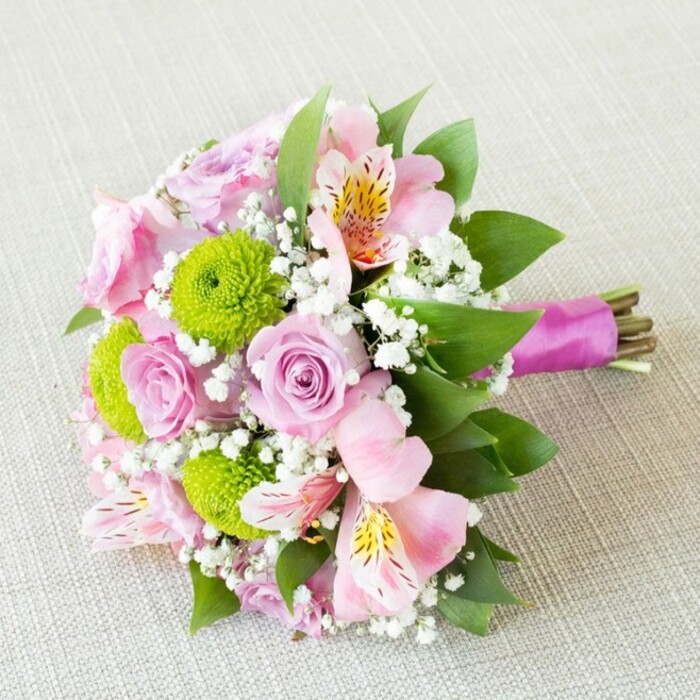 This exquisite, hand-tied bouquet epitomizes the floral artistry found at Brent Cross Flowers, resting on a subtly textured light beige background that complements its pastel and vibrant tones. The bouquet features a seamless interplay of lavender-pink roses with delicate spiraled petals, suggesting a fresh fragrance and creating a soft romantic focal point. Poised among the roses are lively, lime-green button chrysanthemums, their tight clusters lending a modern, sculptural sophistication while brightening the bouquet with youthful energy-an echo of Brent Cross's lively city vibe. Interspersed pale pink alstroemeria, their petals brushed with golden and burgundy flecks, open broadly to invite the eye and recall the diversity of blossoms seen at local flower markets. Wispy white gypsophila (baby's breath) is scattered throughout, imparting a whisper-soft, ethereal finish that lifts the entire arrangement. Rich green leaves frame the composition, grounding the blooms and delivering a sense of fullness and lushness reflective of nearby parks. The stems are neatly gathered and secured by a glossy magenta satin ribbon, tied for style and comfort, and finished to expose the bouquet's vibrant structure. This piece is perfectly suited as a bridesmaid bouquet for a Brent Cross wedding, as a centerpiece for an anniversary, or a fresh gift for homes near Golders Green, exuding feelings of happiness, springtime renewal, and timeless elegance in North London.