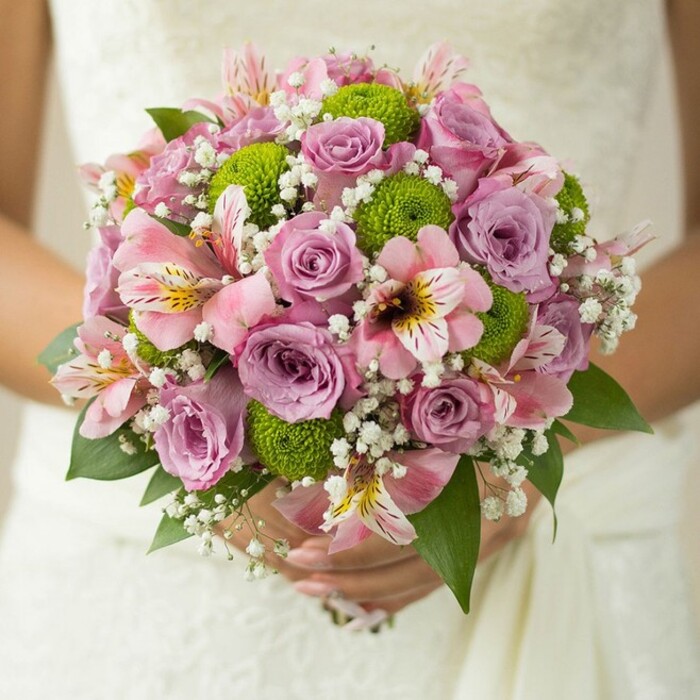 A vision of elegance and joy, this bridal bouquet radiates with the fresh charm of Brent Cross's local artistry. Gently held against the intricate white lace of a wedding dress, the bouquet's rounded form is a canvas for softly layered roses in light pink and gentle lavender, their petals edged with droplets that glisten like morning dew gathered in Brent Park. Adding a spark of contemporary colour, button chrysanthemums in vivid lime green nestle between the roses, their playful, spherical shapes reminiscent of garden play in nearby Claremont Park. Delicately marked pink alstroemeria lilies add both texture and intrigue, their petals veined with maroon and yellow tracing a dance through the bouquet. Filling in each interval, ethereal sprigs of white baby's breath impart volume, softness, and a sense of floating romance-like catching glimpses of white blossoms along the River Brent. Framed by polished dark green leaves, the arrangement is not only tactile but visually stunning, captured in a close-up that reveals every carefully placed petal and stem. This bouquet is the very essence of a Brent Cross wedding: fresh, romantic, and thoughtfully composed for a joyful celebration that honors both tradition and modern flair, perfect for any North West London event.