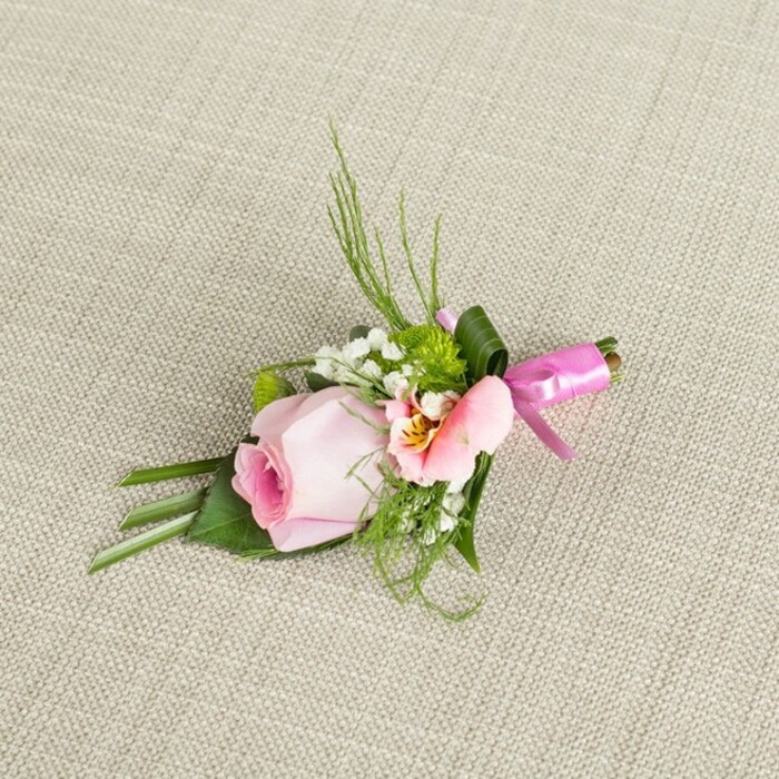 Diagonally placed on an ecru-hued, gently textured cloth reflecting the harmonious calm of Brent Cross, this elegant boutonniere features a luxurious blend of colour, texture, and form. A single blush pink rosebud stands as the arrangement's heart, every petal tightly furled, exuding freshness and promise-emblematic of romantic moments shared in the area's quiet crescent parks. Surrounding the rose, a pale pastel alstroemeria blooms with peach and golden accents, delicate streaks near its centre adding visual intrigue. Wispy sprays of pure white baby's breath evoke a sense of softness and celebration, while playful green button chrysanthemums infuse a cheerful, contemporary twist. Varied foliage-sleek emerald grass blades and darker, oval-shaped leaves, some stylishly folded-provide volume and a lush backdrop, echoing the grace of local woodlands and gardens. The entire cluster is bound at its base by a lustrous fuchsia pink satin ribbon, tied in a petite, elegant bow with careful regard for detail, its sheen adding festive joy and marking the work of Brent Cross Flowers' skilled hands. The boutonniere's restful angle and lighting highlight its balanced arrangement, capturing the gentle emotion and expert design that make it a fitting adornment for weddings and celebrations throughout NW11. Each element whispers of love, possibility, and the joyful artistry of Brent Cross's flourishing floral scene.