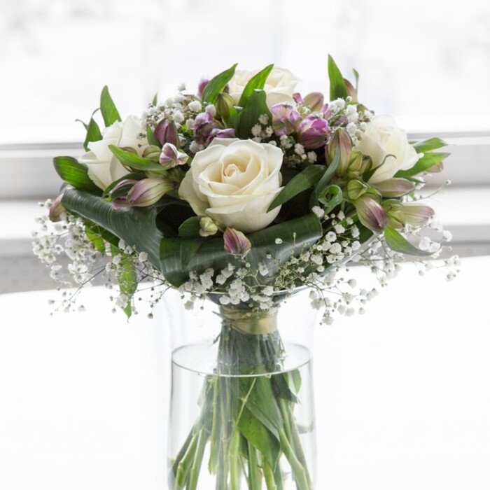In this image, a classic clear glass cylinder vase cradles an abundant bouquet of white roses, lavender alstroemeria, and fine sprays of baby's breath, all bathed in a soft, diffused light that suggests a bright but gentle Brent Cross morning. The roses, positioned as the central focal blooms, are fully opened yet still pristine, their creamy petals layered in precise spirals that form plush, rounded cups. Each bloom seems to invite touch, with a velvety surface that visually conveys softness and quiet romance. Encircling the roses, clusters of alstroemeria in muted lilac and cool purple tones introduce subtle colour and intricate patterning. Some flowers are half open, revealing their inner throats etched with darker streaks and freckles, while others remain in elongated buds, sheathed in green, hinting at unfolding beauty over the days to come. Scattered throughout the design, tiny blossoms of baby's breath create a gossamer haze, their minuscule white flowers perched on slender, branching stems like little drops of light suspended in the air. Lush green foliage frames the arrangement: broad, shining leaves provide a deep green backdrop, while finer, more linear greens add lift and texture, helping to sculpt the bouquet into a graceful, rounded silhouette. Below, the stems are clearly visible through the transparent vase, trimmed to a neat, even length and standing in clear water, a detail that speaks to the professionalism and care you might expect from a trusted Brent Cross florist preparing same-day deliveries to nearby Finchley Road and Temple Fortune. The softly blurred, pale background keeps all attention on the flowers, enhancing their serene character. The overall impression is elegant yet comforting-a bouquet equally suited to offering sympathy, celebrating a quiet anniversary dinner near Clitterhouse Playing Fields, or simply brightening a hallway with its gentle promise of peace, renewal, and enduring affection.
