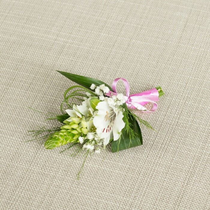A beautifully composed boutonniere is displayed against a softly textured, light beige fabric backdrop, whose linen-like weave brings a warm, natural feel and allows the floral details to stand out clearly. Centre stage is a pristine white alstroemeria blossom, fully open and gently cupped, its broad petals edged with a delicate ripple and marked near the throat by fine reddish-brown spots and streaks that draw the eye inward. Beside this central bloom sits a striking lime-green bud cluster shaped like a tapered cone, similar to an Ornithogalum spike, each tiny, unopened bud pressed neatly against the next to create an intriguing architectural element. Draped around these focal flowers are airy threads of classic white baby's breath, the tiny round blossoms hovering like a light mist, softening edges and lending an almost cloud-like frame to the composition. A base of deep, glossy green leaves supports the flowers, their pointed, blade-like shapes arranged in layers, while slender, bright green grass-like strands twist and arc outward, adding movement and texture that mimic the natural, slightly windswept greenery you might notice walking near Brent Cross's open spaces. To the right, the stems are gathered and meticulously bound with a vivid pink satin ribbon, fashioned into a small, symmetrical bow whose smooth, reflective surface contrasts beautifully with the matte petals and the woven background. The lighting is even and gentle, highlighting every nuance: from the soft translucence of the alstroemeria petals and the tight smoothness of the buds to the gossamer quality of the baby's breath. The overall mood is refined yet joyful-a handcrafted floral accent ideal for a wedding day, prom night, or anniversary dinner in and around Brent Cross, where such details quietly elevate a special outfit and mark the significance of the occasion.