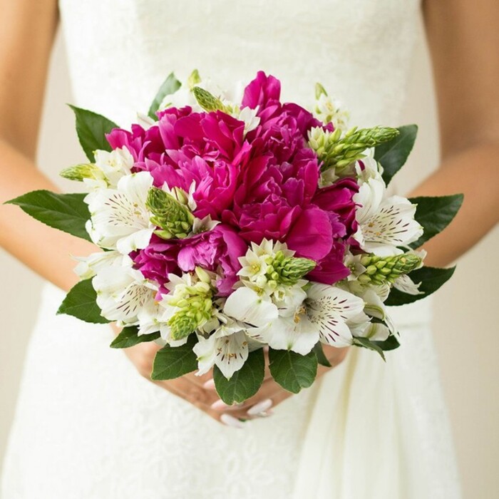 The photograph captures a bride from the waist down, her torso framed by a soft, off-white wedding gown while she holds a rounded bouquet that glows with magenta and white in the centre of the frame. Dominating the arrangement are full, deeply ruffled peonies in intense fuchsia tones, each bloom packed with layers of petals that curl and twist inward, creating shadows and highlights that suggest both softness and depth. Tucked between these bold, central flowers are white alstroemeria, their more slender petals opening gracefully around throats delicately flecked with maroon and fine striping, lending a sense of refined detail when you look closely. Rising up through the mix, spiky, elongated green buds-likely Ornithogalum or similar-add a modern, architectural feel; their crisp lime-green colour injects a lively brightness that prevents the bouquet from feeling too heavy. Encircling the flowers, wide, dark green leaves form a tidy collar, their glossy surfaces catching subtle reflections and helping define the bouquet's rounded silhouette. The bride's hands appear at the base, fingers wrapped gently around the bound stems, nails painted a soft, bridal pink that quietly echoes the feminine tones of the blooms. The background is blurred to a gentle haze, focusing attention on the interplay of colour and texture in the flowers and hinting at lace detail on the dress without pulling focus. The overall impression is of a carefully composed bouquet that might have been carried out of a Brent Cross ceremony room, ready for photos in a nearby park or garden square, its fragrance mingling with the cool London air. It feels both classic and fresh, like something chosen by a bride who wanted her flowers to express joy and personality while still looking timeless in her wedding album.