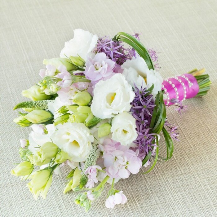 Lying diagonally across a softly hued, textured fabric-evocative of quality linens at Brent Cross's local venues-this hand-tied bouquet sweeps the eye with its delicate, harmonious palette. At its core, several large white Lisianthus blossoms open fully, their soft yellow-green centers and ruffled petals bringing a radiant purity. These are seamlessly accented by slender lisianthus buds in pale lime and white, hinting at graceful growth. Pastel lavender-pink stock flowers cluster nearby, infusing soft texture and a gentle sweetness, reminiscent of early blooms seen along Brent Terrace gardens. Deep purple allium flowers burst upward, their star-shaped clusters making a bold statement and providing a sophisticated, architectural flair. Wispy green filler elements weave delicately between blossoms, while arcs of fresh bear grass loop through the bouquet, giving it structure and gentle dynamism. At the base, stems are tightly wrapped in vibrant fuchsia satin ribbon, elegantly finished with a luminous white pearl strand-a refined touch echoing timeless Brent Cross festivities. Illuminated by diffused daylight, every hue stands out in invitation: creamy whites, pastel lavenders, and bold purples, all united in an arrangement that channels celebration, local artistry, and contemporary romantic style-an ideal accompaniment for any Brent Cross wedding or milestone event.
