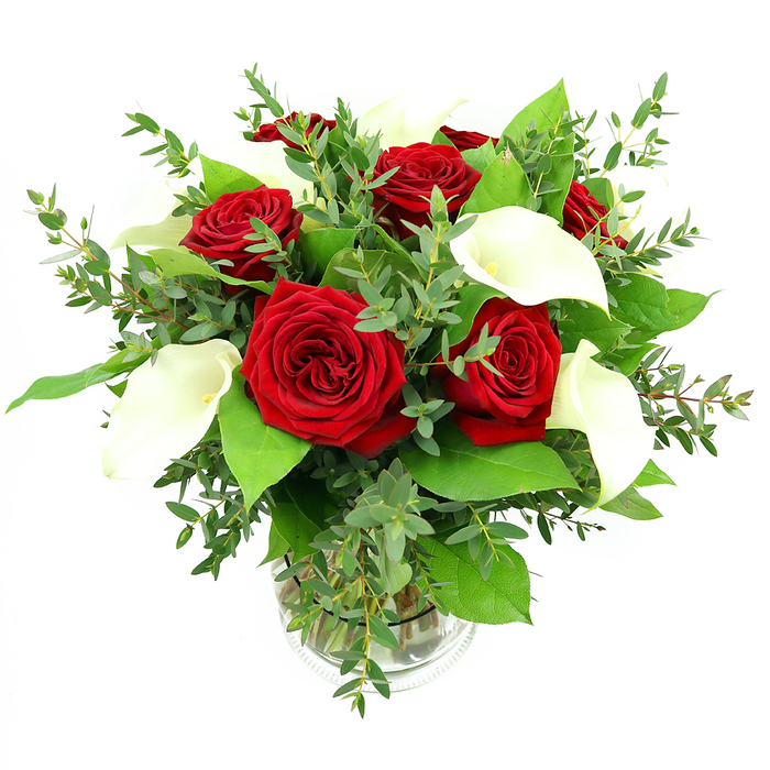 A beautifully balanced bouquet of deep red roses and white calla lilies is displayed in a clear, cylindrical glass vase, photographed against a clean white background that amplifies the vibrancy of the flowers. At its heart, roughly six to seven roses are in full bloom, their velvety petals arranged in intricate spirals of rich crimson that give each flower an opulent, almost plush appearance. Between them stand four or five calla lilies, their tall, trumpet-shaped spathes of pure white tapering to elegant curves at the rim, with slim yellow-orange spadices visible in the centre, adding warmth and dimension. Surrounding and supporting these focal blooms is an abundant mix of greenery: airy eucalyptus stems with slender, rounded leaves in a silvery-blue-green tone trail softly around the outer edges, bringing gentle movement, while broader, darker green foliage anchors the arrangement with a deep, glossy backdrop. The stems are neatly aligned within the transparent vase, some crossing delicately, and are clearly visible through the water, while a fine ribbed band near the base of the vase introduces a subtle textural detail. The whole composition is bathed in bright, even lighting, which enhances the contrast between the crimson roses, the luminous white lilies, and the layered greens, highlighting the freshness of every petal and leaf while maintaining a serene, uncluttered feel. This is the kind of arrangement you might order from Brent Cross Flowers for an elegant anniversary dinner in a flat just off the North Circular, a romantic surprise delivered to a home near Brent Cross Shopping Centre, or a sophisticated table centerpiece for a gathering in nearby Cricklewood or Temple Fortune.