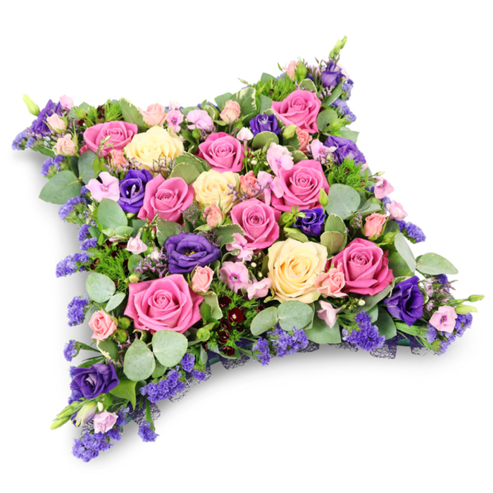 The photograph shows a sumptuous floral arrangement in the form of a cushion, a traditional pillow-shaped funeral tribute that looks as though it has been gently laid down in stillness. The cushion itself is a soft rectangle with rounded corners, entirely concealed beneath dense layers of roses, lisianthus, and fine filler flowers. The overall palette is composed of romantic pinks, warming creams and pale yellows, and striking deep purples. Large, full-headed roses form the backbone of the design: some are bright fuchsia, others are delicate shell pink, and others yet are a creamy, buttery yellow, each head placed snugly beside the next until the surface resembles an intricate patchwork of petals. Rich violet-purple lisianthus blooms are threaded throughout, their gently ruffled forms adding movement and a plush, velvety feel that contrasts with the more structured roses. Smaller spray roses in soft pink are tucked into gaps, while tiny clusters of lavender-blue filler flowers, similar to statice or limonium, bring a haze of colour and an almost lace-like texture across the pillow's face. Around the edges, and peeking between blossoms, foliage in mixed greens anchors the colours: rounded, silvery eucalyptus leaves lend a cool, misty tone, and darker, fine-textured, fern-like greenery gives depth and a natural frame. The arrangement is photographed against a crisp white backdrop, a faint shadow beneath accentuating the pillow's gentle height and giving the impression of quiet presence. It is easy to imagine this floral cushion being delivered for a service near Brent Cross Shopping Centre or a nearby cemetery, chosen by a family who want to honour their loved one with a tribute that communicates tenderness, remembrance, and the wish that they might rest on a bed of soft, enduring beauty.