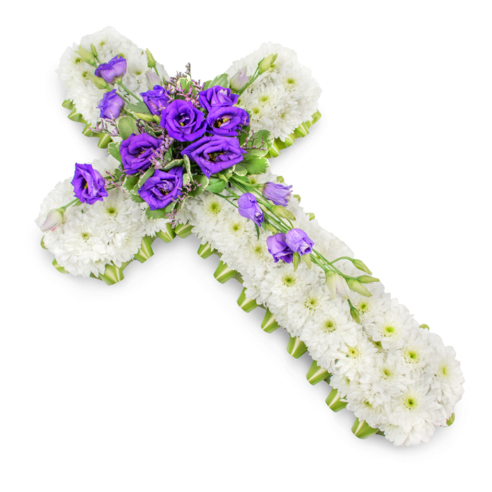In this carefully lit photograph, a large floral cross lies against a pure white backdrop, allowing its serene colours and fine craftsmanship to stand out clearly. The cross is entirely covered in white spray chrysanthemums, placed so densely that they form an unbroken blanket of soft petals. Each tiny flower is fresh and full, its pale green centre peeping through and creating a quiet, speckled pattern across the surface, like small points of light in a peaceful sky. A narrow band of lime-green ribbon traces the entire outline of the cross, its crisp edges giving definition to the form and a contemporary freshness that harmonises beautifully with the natural whites and greens of the flowers. At the point where the arms and stem of the cross intersect, a lush, rounded grouping of deep purple lisianthus blooms forms the heart of the design. The blossoms vary from fully open cups, revealing velvety inner petals, to more tightly furled buds that promise to open in time. Subtle foliage in gentle greens, along with a few light filler flowers, is threaded between the lisianthus, lending natural movement and softly blurring the transition between the central spray and the chrysanthemum base. From this focal cluster, a delicate extension of lisianthus stems sweeps diagonally down the lower right side of the cross, their blossoms and buds forming a loose cascade that feels both elegant and unforced. A very soft shadow beneath the arrangement adds depth without disturbing its tranquil mood. This reverent floral cross reflects the kind of tributes chosen by families across Brent Cross, Cricklewood, and Finchley, perfectly suited to a church altar, crematorium chapel, or graveside, where it serves as a gentle expression of faith, gratitude, and remembrance.