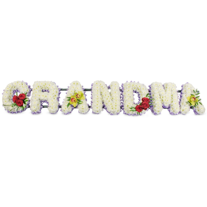 The photograph shows a long floral display spelling out GRANDMA in carefully shaped capital letters, presented against an uncluttered white background that keeps the focus on the flowers themselves. Each letter is expertly covered in small white chrysanthemums or similar blooms, packed tightly so that the surface appears smooth and plush, suggesting both softness and formality. Beneath this white blanket, a narrow edging of pale lavender or soft purple flowers gently frames each letter, adding definition without overwhelming the purity of the main colour. On the G, placed toward the left, three deep red roses rest at the lower left curve, their petals rich and velvety, emerging from a nest of glossy dark green foliage that adds depth and structure. The first A carries two yellow-green orchids arranged along its crossbar, each orchid showing a distinctive reddish-brown centre that draws the eye and hints at a more personal, thoughtful touch. The letters R, N, and D stand quietly in the middle, pure white with their lavender halo, unadorned by additional colours so that the tribute retains a sense of peaceful continuity. Moving further right, the M holds another trio of red roses in its lower right segment, visually balancing the red cluster on the G, while the final A repeats the placement of two yellow-green orchids, closing the word with the same gentle brightness that appears near the start. All the letters are attached to a slim dark metal frame that sits almost invisibly behind them, supporting the tribute for display at funeral services, cemeteries, or crematoria around Brent Cross, Staples Corner, and the nearby North Circular. The overall expression is one of respect, affection, and quiet remembrance, a fitting floral salute to a much-loved grandmother at the heart of the family.