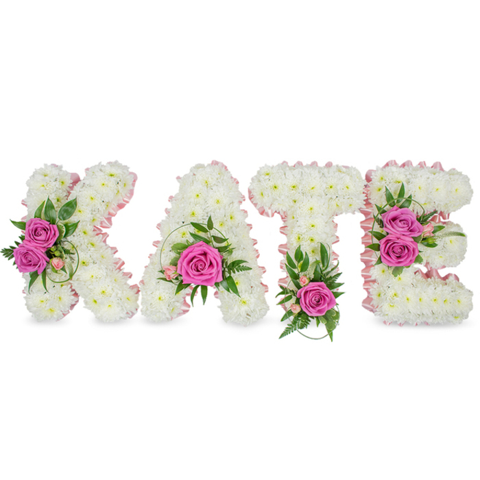 A striking, custom-made floral name tribute spells KATE in large, three-dimensional block letters, each letter built from hundreds of small, pristine white chrysanthemums to form a soft, cloud-like mass. The chrysanthemums are packed tightly, their rounded petals creating a seamless, pillowy surface that holds shape while suggesting a delicate touch. A soft pale-pink satin ribbon has been expertly applied to outline the inner contours of every letter, adding a refined, feminine border that catches highlights and gives the letters a finished, ceremonial quality. Hot pink roses are used as focal accents: the K carries two open rose blooms on its left arm amidst a spray of small pink buds and glossy green leaves; the A shows a single prominent rose nestled low in its triangle with slender green sprigs; the T is balanced by one vivid pink rose at its base with fern-like foliage; and the E is brightened by two roses on its right side accompanied by variegated leaves and fine pink floral details. Set against a pure white background with subtle shadows beneath, the composition reads as a freestanding memorial display - a bespoke piece often requested for funerals, memorial services, or intimate celebrations of life. The faint scent of roses and the tactile variation between chrysanthemum fluff and the smooth rose petals lend sensory depth. Crafted with local knowledge from Brent Cross Flowers and suited for delivery throughout Brent Cross, Hendon, and nearby neighbourhoods, this tribute communicates respect, love, and the careful hand of a florist experienced in personalised sympathy work.