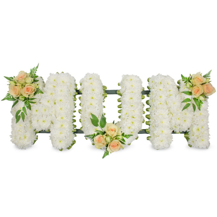A carefully arranged floral tribute spells out MOM in large, elegant block letters, photographed against a spotless white background that accentuates the quality and care of the floristry. The three letters are mounted on an unseen supportive frame and generously padded with a dense covering of pure white flowers, likely small-headed chrysanthemums or spray carnations, each individual bloom pressed tightly beside the next. This creates a soft, quilted surface that is almost touchable, conveying an air of tranquility and respect well-suited to a funeral or memorial service in the Brent Cross area. A narrow band of dark green ribbon traces the outer edges of the letters, and the frame itself peeks through just enough to provide definition and structure, much as a local florist from Brent Cross Flowers would ensure for safe delivery to a nearby church or crematorium.   Adding warmth and a sense of personal story, three rounded clusters of peach and apricot-coloured spray roses are placed strategically along the word. One rests near the upper left of the first M, one nestles into the lower central point of the O, and the final cluster is cradled at the upper right of the last M, drawing the eye gently from left to right. The roses themselves are at different stages of opening, with tight buds, half-open blooms, and lush, fully opened flowers all present, offering a subtle suggestion of different moments in a life. Each grouping is cushioned by sprigs of vibrant green foliage, from lacy fern-like leaves to stronger, glossy greens that add depth, contrast, and a touch of garden freshness. The overall arrangement feels both classic and personal, an eloquent floral message of love, remembrance, and gratitude for a mother, suitable for funeral services across Brent Cross, Hendon, and nearby Temple Fortune.