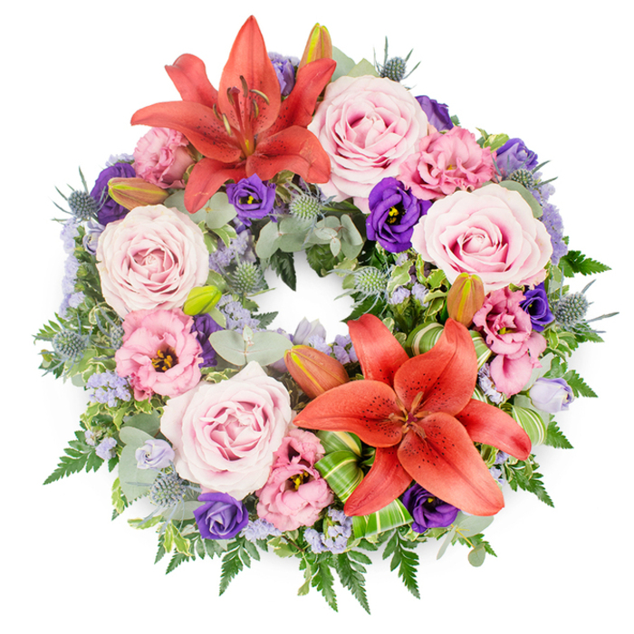Captured against a bright white background, this full sympathy wreath forms a perfect circle of colour and foliage, designed with the care of an experienced local florist. The structure begins with a dense collar of greenery: rounded, almost matte eucalyptus-style leaves, fine feathery fronds, and clusters of darker green foliage weave together to create depth and ensure there are no gaps, so the ring appears lush from every angle. On top of this verdant base, two vivid coral-orange Asiatic lilies serve as the primary focal points. One sits near the upper centre of the wreath, while the other rests slightly lower, so the eye naturally travels between them. Their wide, star-shaped petals curve backward to reveal tall stamens, giving a sense of openness and quiet vitality. Between and around these lilies, roses in soft blush and lavender-pink nestle closely, their spiral centres gently unfolding as if caught at the perfect moment between bud and full bloom. Alongside them, eustoma (lisianthus) flowers in pale pink and deep purple provide a ruffled contrast, filling small spaces and adding richness through their cup-shaped, layered petals. Dotting the design, small spiky heads of blue-grey eryngium rise from the greenery, bringing a cool, slightly metallic tone that balances the warmth of the lilies and roses. Threads of tiny lavender-blue filler flowers wind lightly through the arrangement, forming a delicate veil that softens the transitions between blooms and foliage. A few closed or partially open lily buds peek from the leaves, hinting at new blossoms yet to come. Near the lower lily, carefully folded bright green leaves, likely aspidistra or ti, add a contemporary, structured detail that signals professional craftsmanship, the sort you might find at Brent Cross Flowers for services held near Hendon Way or throughout the wider Brent Cross community. Overall, the wreath conveys peaceful remembrance and enduring affection, an appropriate tribute for a funeral or memorial gathering.