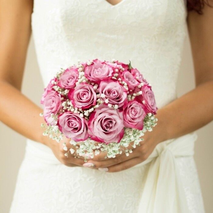 Framed by the pure elegance of a white, sleeveless wedding gown, this intimate photo focuses on a bride-or guest of honour-standing amidst the nostalgia of Brent Cross, cradling a compact, round bouquet close to her heart. The bouquet is a masterwork of about twelve roses, each displaying a gentle gradient from a tender, milky-pink core outward to show-stopping fuchsia and magenta petal tips, providing an impression of movement and fullness. Interlaced between the roses, dozens of tiny, snowy white baby's breath blossoms lend an airy, dreamy texture reminiscent of celebratory mornings beside Brent Cross parks. Subtle hints of green peek out, barely visible but ensuring the sense of a living, freshly gathered design. The hands holding the bouquet-soft, with tastefully painted nude nails-radiate grace, while the flowing lace and sash of the gown provide layered interest and classic British bridal charm. The neutral, softly blurred background turns the spotlight onto this floral treasure, evoking the meticulous attention to detail provided by local florists. This vision captures the enduring romance and emotional significance of wedding bouquets, embodying the joy and anticipation of North West London's cherished marriage celebrations.