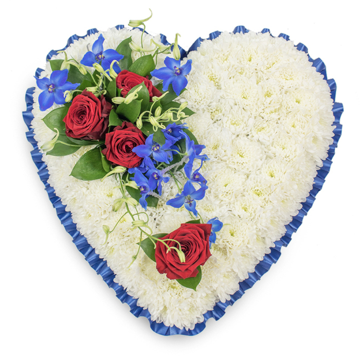 A large, heart-shaped floral tribute is the sole focus of this photograph, captured from directly above to highlight its sculpted form and meticulous design. The heart's interior is completely filled with white chrysanthemums, their numerous, delicate petals overlapping in dense layers so that the surface appears soft and cushion-like. Every blossom is clean and bright, contributing to an atmosphere of calm and purity. Framing the outer edge, a royal blue satin ribbon has been pleated into gentle ripples, forming a ruffled border that contours smoothly around the heart. The ribbon's glossy texture reflects tiny flashes of light, adding sophistication and depth to the otherwise matte petals. On the upper left portion, an expressive arrangement of feature blooms sits proudly atop the chrysanthemum bed: three deep red roses, fully open, reveal spirals of velvety petals. They are supported by structured dark green foliage, which gives a grounding framework and emphasises the roses' richness. Extending from this cluster, slender stems of electric blue dendrobium orchids trail diagonally towards the centre, their star-shaped petals blending vivid blues and teals to create a striking contrast against the white. Pale green curly tendrils interlace with the orchids and roses, softening edges and hinting at natural movement. On the lower right curve of the heart, an isolated red rose rests gently on the white background, accompanied by just a whisper of greenery. This single bloom mirrors the main cluster's deep hue while conveying a quieter, more personal note of farewell. Overall, the interplay of white, red, and blue feels both dignified and heartfelt, the kind of memorial heart a family in Brent Cross might select to honour a loved one, whether for a funeral at a local synagogue, church, or crematorium near the North Circular.