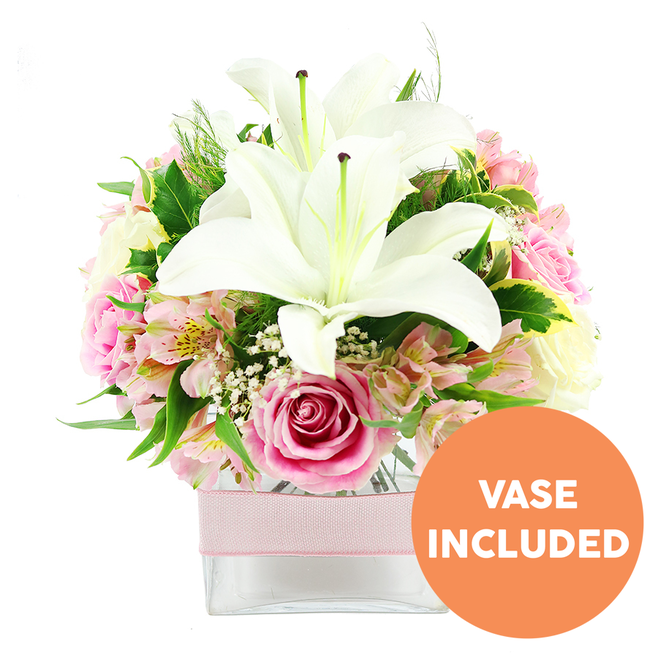 A lush, romantic floral arrangement is captured against a bright white backdrop, conjuring the atmosphere of a calm, uncluttered room in Brent Cross. At the heart of this design, two large white Oriental lilies are fully open, their petals thick, smooth, and slightly reflexed, creating a wide, star-shaped form. Dark reddish-brown stamens rise from the centre of each lily, dusted with pollen and providing a striking contrast against the creamy white. Surrounding these lilies, a generous assortment of pink roses is arranged in a harmonious dome, their petals at varying stages of unfurling-from tight buds that hint at colour to lush, fully open blooms with layered, ruffled centres that evoke softness and warmth. Between the roses and lilies, slender stems of alstroemeria in blush pink and white weave delicately, their fine petals and subtle yellow speckling adding detail and a slightly whimsical charm. Interlaced throughout are sprays of white baby's breath, each tiny flower forming part of a cloud-like cluster that softens the transitions between blooms and gives the piece a light, airy quality. A mixture of green foliage frames the composition, combining broad, glossy leaves with finer, feathery greenery that evokes the leafy edges of nearby Golders Hill Park, enhancing the freshness and sense of natural abundance. All of this is presented in a low, square clear glass vase, the stems neatly visible through the transparent sides and resting in clear water. Around the centre of the vase, a pale pink ribbon is wrapped neatly, lying flat and unknotted, introducing a subtle, elegant band of colour. In the bottom right corner of the image, an orange circle with bold white capital letters clearly states VASE INCLUDED, emphasising that this bouquet arrives complete and ready to display, suitable for birthdays, anniversaries, or simply sending a touch of beauty to someone living near Brent Cross Shopping Centre.