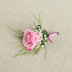 An elegant pink rose boutonniere lies gently on a natural, light beige linen background, the woven threads of the fabric creating a calm, neutral stage reminiscent of a carefully laid wedding suit on a bedroom chair near Brent Cross station. The focal rose is semi-open, its velvety petals forming a soft cup that gradually unfurls toward the edges, where the pink deepens into a slightly more vibrant, romantic shade. Around the base of the rose, tiny white baby's breath blossoms cluster in airy sprays, their cloud-like texture lending a classic bridal feel and enhancing the sense of lightness. Fine, grass-like green foliage arcs out from behind the bloom, giving the arrangement a gentle sense of movement, as if caught in a breeze along the paths by Brent Cross Shopping Centre. A single, broader green leaf is carefully folded and wrapped to cradle the base, adding structure and a clean, tailored look to the small floral piece. The stems are bound with a lustrous satin ribbon in harmonising tones of lavender-pink and deeper magenta, wrapped smoothly and tied into a modest knot that keeps the focus on the rose while adding a subtle hint of colour-coordinated luxury. Soft, diffuse lighting falls evenly across the image, highlighting the petal gradation, the crisp edges of the greenery, and the natural, matte texture of the linen beneath. This boutonniere feels perfect for a groom or prom guest heading out from a Brent Cross home to a ceremony in nearby parks or venues, carrying with them a small, handcrafted symbol of romance and celebration made with the care of an experienced local florist.