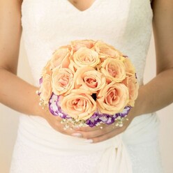 The image presents an intimate, waist-up view of a bride, her figure centred against a light, almost glowing backdrop that could just as easily be a bright room off Hendon Way as a sunlit bridal suite near Brent Cross. Her wedding dress is a timeless white, the bodice ornamented with fine lace motifs that trace along a soft V-neck and down towards a gently cinched waist, where smooth, layered fabric falls into a flowing skirt. Her hands are positioned just below her midline, fingers loosely interlaced around a bridal bouquet that is clearly the focal point of the photograph. The bouquet is round and compact, meticulously styled so that each flower sits perfectly in place. The upper portion is composed of many plump roses in shades of peach, light orange, and blush pink; the colours blend like a muted sunset, with outer petals paler and centres deepening into warmer, romantic tones. The roses' velvety, tightly packed petals create a sense of fullness and luxury, as though each bloom has just been chosen from a fresh delivery that morning. Encircling this rose cloud is a dramatic band of purple blooms with ruffled petals tipped in white, likely lisianthus, offering striking contrast and a slight swirl of movement where their edges curl. Around the base, delicate clusters of white baby's breath form a soft, cloud-like fringe, echoing the bride's lace and adding airiness to the tight structure. The quiet sheen of her nude-toned manicure, with one subtly glittered thumb, introduces a personal, modern detail. The scene feels calm, romantic, and carefully composed, like a cherished wedding portrait taken moments before leaving a Brent Cross home to head to the ceremony.