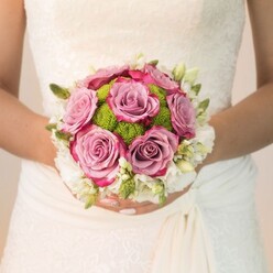 Lit by soft daylight streaming through a Brent Cross window, a bride's delicately patterned ivory gown provides a stunning canvas for her artfully arranged bouquet. The rounded design draws the eye to its heart, where plush, full-bloom roses unfurl in shades of mauve, lavender-pink, and fuchsia-their elegant transitions embodying both softness and spirited celebration. Encircling the roses are vibrant button chrysanthemums in zesty green, their unique, dewy textures providing contrast and a burst of cheerful energy, much like the vibrant spirit of Brent Cross's high streets and gentle parks. Completing the composition, a border of white blooms-sprightly freesia or petite spray roses-offers an ethereal touch, accented by slender green buds and leaves that give depth and freshness. The bride's hands, beautifully manicured in light pink with a subtle shimmer, gently cradle the bouquet, reflecting the poise and anticipation of the moment. Every bloom appears meticulously selected by Brent Cross's talented local florists, whose experience shines in the bouquet's harmonious palette and refined balance. This image eloquently captures the artistry and heartfelt joy woven into local bridal traditions, ideal for weddings held throughout Hendon, Golders Green, and NW2.