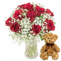 This image captures a tender, romantic gift pairing, photographed in a clean, contemporary style that keeps every element in sharp focus. On the left, a clear glass vase sits firmly on a white surface, its cylindrical shape allowing the viewer to see the straight, neatly trimmed stems of the flowers inside. The water level is visible and crystal clear, suggesting freshness and careful preparation, much like the hand-crafted bouquets made for same-day delivery around Brent Cross. Rising above the rim of the vase is an abundant bouquet of approximately one dozen red roses. Each bloom is full and lush, with velvety petals unfurling from tightly packed cores into wider, more relaxed edges that form a classic spiral shape. The colour is a rich, romantic crimson, deepening to a burgundy tone in the shadowed inner layers. Between these roses weave many slender stems of baby's breath (Gypsophila), each topped with delicate clusters of tiny white flowers. These airy blossoms create a gentle halo effect around the roses, softening the intensity of the red and adding a dreamy, cloud-like texture. Occasional flashes of dark green foliage peek out from behind the flowers, giving the arrangement fullness and a natural garden feel. To the right of the vase, a plush teddy bear sits upright, facing forward as if waiting to be picked up. Its fur is a soft, light brown, appearing fluffy and inviting to the touch. The bear has classic features: small black button eyes that lend a sweet, attentive gaze, a neatly stitched nose, and a subtle embroidered smile. A dark brown ribbon is tied in a simple bow at its neck, adding a touch of sophistication. Set against a pure white background, the combination of vivid red roses, floating white gypsophila, and the comforting teddy bear evokes emotions of love, comfort, and celebration-perfect for marking anniversaries, Valentine's surprises, or heartfelt birthdays across Brent Cross and nearby areas of North London.