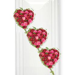 This image shows a striking triple-heart funeral tribute, created entirely from roses and ivy, suspended against a luminous white background. Three heart-shaped arrangements are stacked in a gentle diagonal, starting at the top left and gently cascading down to the bottom right, like a visual echo of love repeated and remembered. Each heart is filled to the edges with roses in shades of deep crimson, bright fuchsia, and soft pink, the blooms nestled so tightly together that they form a lush, almost cushioned surface. The variation in bloom stages-from just-opening buds to fully unfurled roses-adds texture, depth, and a sense of life to the design. Encircling each heart, delicate trails of fresh green ivy trace the contours with care, while longer strands extend downward to link the hearts, suggesting relationships that remain intertwined even after loss. The colour story is both rich and comforting: the dark red roses convey respect and enduring devotion, while the pink roses introduce a tender, affectionate note, ideal for farewell services that celebrate a life full of warmth. The pristine white wall behind the tribute, reminiscent of a quiet chapel or funeral room near Brent Cross Shopping Centre or along Hendon Way, helps the colours stand out with clarity. Gentle lighting creates soft highlights on the petals and barely-there shadows to the right, giving the arrangement a calm, dignified presence. Overall, the design feels like a heartfelt message crafted by an experienced local florist, meant to accompany a final goodbye with beauty, grace, and the reassurance that love remains, even in parting.
