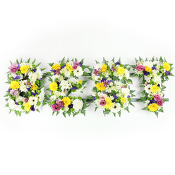 A clean, head-on view shows a horizontal floral lettering tribute spelling BEST, each capital letter densely packed with freshly arranged flowers and edged with rich fern fronds against a pure white backdrop. The overall tone is bright and uplifting: cheerful yellow roses and gerbera daisies dominate alongside generous white chrysanthemums whose many petals create a soft, cloud-like texture. Interspersed through the composition are dusky lavender lisianthus and striking magenta spray roses or carnations that create lively, respectful accents. Tiny clusters of pale green and cream filler blooms nestle between the larger heads, creating gentle pockets of negative space that let the main flowers breathe while adding tactile depth. The fern leaves act as a dark, graceful frame for each letter, their lacy fronds curling slightly over the petals and lending the piece a natural, garden-fresh finish.

A subtle dark green or metal support bar is visible in places - notably within the horizontal arm of the E and near the base of the T - showing the structural care taken to keep the letters upright during procession or display. The photograph's bright, even lighting highlights the freshness of each bloom, the satin sheen on rose petals and the feathery texture of chrysanthemum disks. Scent, though unseen, is suggested: a gentle rose sweetness with a hint of green, comforting rather than overpowering. Thoughtfully composed, this floral word tribute balances dignity and celebration, a fitting choice for memorials or remembrance services, and a familiar piece created for families from Brent Cross through to nearby Cricklewood.