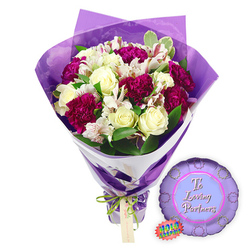 Against a simple white background, this image captures a romantic bouquet and balloon pairing that feels perfect for an anniversary dinner or quiet celebration at home in Brent Cross. The bouquet itself is full and carefully balanced, with creamy white roses at the heart, their petals tightly layered in soft spirals that suggest both purity and devotion. Nestled between them are generous heads of deep magenta and burgundy carnations, their dense, ruffled petals forming plush cushions of colour that bring warmth and drama to the design. Fine stems of white alstroemeria are threaded throughout, each blossom revealing slender inner petals delicately striped and dotted with reddish-brown markings, adding a refined, almost painterly detail that invites closer attention.   

The flowers are cradled by an abundance of fresh green foliage: some leaves are smooth and deep green, while others show subtle creamy edges, giving the bouquet texture and a slightly wild, garden-inspired feel, as though gathered after a walk near Brent Cross Shopping Centre's surrounding green spaces. The arrangement is wrapped first in a crisp white paper layer that provides a clean, structural base and peeks out in a diagonal slash of brightness. Over this, a sheer purple cellophane layer, softly crinkled, sweeps upward and outward, framing the flowers with a translucent amethyst glow and catching highlights of the studio light. At the base, the stems are neatly bound with a bright green satin ribbon tied in a small bow, which also holds a light-brown rectangular gift tag ready for a loving message. To the right, resting on the white surface, is a round metallic purple foil balloon with a scalloped, rosette-like edge. Its centre, in a paler lavender circle, bears the words "To Loving Partners" written in graceful pink script, with a small holographic sticker glinting nearby, together creating a scene that beautifully expresses affection for couples across Brent Cross.