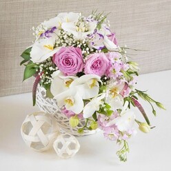 A lush, romantic centerpiece overflows from a crisp white wicker basket, captured in soft, diffused light that gives the scene the calm, polished feel of a Brent Cross wedding reception. At the heart of the arrangement, fully opened pastel pink roses spiral outward in tight, velvety layers, their petals curling like satin ribbons and conveying warmth and affection. Nestled between them, white Phalaenopsis orchids spread their broad, butterfly-like petals, each bloom showcasing a golden yellow heart with fine speckles, introducing an air of luxury often associated with high-end floristry. Surrounding these focal flowers, white Lisianthus blossoms add gentle ruffles and depth, some petals touched with the faintest lavender-purple edge that cools the palette and creates subtle visual interest. Tall stems of pale pink stock weave upward and gently lean outward, their clustered blooms forming soft plumes that add movement, fragrance, and a sense of abundant ease, as if freshly arranged for a family gathering near Brent Cross station. Delicate sprays of baby's breath are scattered through the bouquet, their tiny white flowers floating like a mist, lightening the composition and filling it with a cloudlike softness. Deep green foliage peeks between the petals, with a few slender leaves and buds escaping over the rim of the basket, lending a natural, slightly unstructured charm. The tightly woven white basket sits on a glossy white surface, reminiscent of a laid wedding table or modern kitchen counter, accompanied by two pale woven decorative spheres whose open, lattice-like structure echoes the basket's texture. A soft beige, linen-textured background frames the entire scene in warm neutrality, allowing the pinks, whites, and greens to glow. As a whole, the image conveys refined celebration and gentle romance, exactly the sort of table arrangement a Brent Cross florist might prepare for weddings, engagement parties, or elegant home entertaining.
