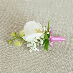 A single, exquisitely composed boutonniere rests on a pale beige, linen-textured fabric, the subtle weave of the material forming a quiet, neutral canvas. At its heart blooms a pristine white Phalaenopsis orchid, the petals broad, velvety, and gently cupped as if holding light. In the centre, soft strokes of buttery yellow and the faintest blush of pink add depth and warmth to the otherwise pure white flower, like a whispered secret at the core of the bloom. Encircling this focal orchid is an airy crown of baby's breath, countless tiny white flowers clustered together to create a cloud-like softness that contrasts with the orchid's smooth, sculptural petals. Slender, glossy green leaves slip through the composition, bringing a touch of fresh foliage that hints at a carefully tended greenhouse or garden. Extending to one side, a short stem carries a pair of tightly closed, lime-green orchid buds, their rounded tips suggesting blooms that will unfurl in days to come, evoking a sense of new beginnings. All the stems are neatly bound by a vivid fuchsia satin ribbon, tied into a small, precise bow, its sheen catching the light to introduce a lively, celebratory flash of colour. The lighting is gentle and even, emphasising the softness of the petals, the feathery detail of the baby's breath, and the tactile quality of the fabric beneath. The design feels intimate and thoughtful, the kind of handcrafted piece a local Brent Cross florist might create for a wedding at a nearby venue, a prom evening, or a special family celebration, carried through the streets around the shopping centre and residential avenues with quiet pride.