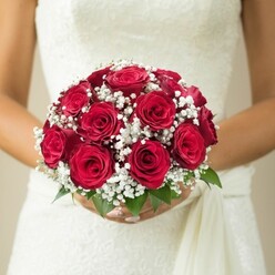 Bathed in soft daylight reflective of Brent Cross's bright mornings, a bride is depicted holding a classic round bouquet of vibrant red roses, their petals plush and full, radiating the warmth of local craftsmanship. The tightly clustered blooms are interlaced with airy sprigs of white baby's breath, their tiny, star-shaped flowers lending subtle movement and contrast-a floral constellation inspired by the tranquil paths of Brent Reservoir. A few vivid green leaves peek out beneath the bouquet, grounding the arrangement in the fresh greenery renowned in the neighbourhood. The bride's delicate hands, manicured and steady, encircle the stems with care, highlighting the ceremony's significance and the gentle artistry behind each arrangement crafted at Brent Cross Flowers. The upper edge of her gown shimmers with textured lace, perhaps echoing design elements seen in elegant wedding venues along Hendon Way. On her side, a soft drape completes the harmonious look. A muted, creamy background lets the bouquet take centre stage, imbuing the image with a sense of hope, calm, and anticipation that defines the most heartfelt North London weddings and inspires those planning their floral moments close to home.