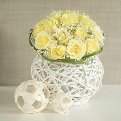 A sophisticated floral centerpiece takes the spotlight, built around a perfectly rounded bouquet of creamy yellow roses that cluster together in a seamless dome. Each rose is in full bloom, petals layered in generous spirals that give a feeling of depth and quiet luxury, while fine sprays of white baby's breath slip between the blossoms, adding a filigree of tiny blooms that lighten the palette and bring a romantic, wedding-day softness. Encircling the base of the bouquet, a narrow band of vivid green leaves has been arranged into a smooth, continuous collar, creating a sharp, fresh edge where the flowers meet the vessel. The arrangement is cradled in a distinctive white vase formed from interwoven strands reminiscent of rattan or fine wicker; the open structure allows light to pass through and cast delicate, lacy shadows on the pristine white surface beneath, giving the piece a modern decorative feel. Resting on the same tabletop are two light-toned woven spheres, one slightly larger than the other, their open latticework echoing the vase's design and completing the sense of carefully curated styling, much like you might see on a beautifully dressed wedding table in a Brent Cross hotel or restaurant. The background is gently defocused in soft beige and cream tones, with no sharp edges to distract from the floral work, which remains the clear focal point. The overall mood is one of calm celebration and refined taste, as though the arrangement has been placed to greet guests at an intimate reception or family gathering, its pale colours and thoughtful details speaking quietly of care and craftsmanship.