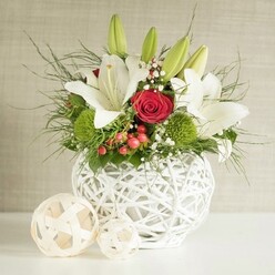 This image presents a meticulously arranged floral centrepiece that would sit beautifully along a wedding banquet table in Brent Cross, perhaps in a boutique hotel or a private dining room just off the North Circular. At the heart of the composition stands a round, sculptural vase in crisp white, fashioned from interlaced strands that form a light, open-weave globe. The texture of the vase, reminiscent of woven wicker yet distinctly contemporary, adds visual interest even before the flowers are seen. From this vessel emerges a generous bouquet dominated by serene white lilies, romantic red roses, bright green pompon blooms, and a cascade of greenery. Several lilies are in full, open bloom, their satin-like petals gently curving backwards to reveal subtle freckles and golden stamens, while a few long buds remain tightly closed, lifting the eye upward and suggesting days of enjoyment ahead. Tucked among these lilies, velvety red roses offer a dramatic burst of colour; one rose, in particular, sits close to the centre, its tightly layered petals forming a perfect spiral that immediately draws attention. Rounded, apple-green dianthus or chrysanthemum pompons introduce a textured, cushiony element that contrasts with the sleek, architectural lines of the lilies. Sprays of delicate white baby's breath weave through the arrangement like a fine mist, softening the overall look, while small clusters of red berries nestle among the foliage, adding a festive, celebratory touch. Wispy asparagus fern fronds and deeper green leaves extend beyond the rim of the vase, giving the bouquet a natural, slightly untamed movement, as though it had just been gathered from a well-tended garden in nearby Hendon or Finchley. To the left of the vase, two pale woven decorative balls rest side by side, gently mirroring the vase's texture and completing a sophisticated, harmonious scene perfect for both weddings and special family gatherings around Brent Cross.