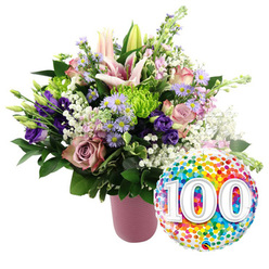 A joyful 100-year celebration arrangement is showcased on a pristine white background, pairing a luxurious bouquet with a bright, confetti-patterned balloon that feels ready to be delivered to a home in Brent Cross. The flowers are artfully arranged in a contemporary cylindrical vase finished in matte dusty pink, with vertical ribbing that adds a pleasing touch of texture. Rising gracefully from the vase, pale pink Oriental lilies take the spotlight, their star-shaped blooms fully open in some places and still partly closed in others, hinting at fragrance and beauty yet to unfold. Around these lilies, an array of roses in soft blush and deeper mauve tones spread their velvety petals in gentle spirals, echoing the passage of years with each unfolding layer. Deep purple lisianthus stems weave in and out of the roses, their slightly ruffled petals adding depth and richness to the pastel palette. Scattered among them, petite lavender asters with golden-yellow centres lend a field-fresh charm, reminiscent of wildflowers near the quieter streets around Brent Cross. To energise the arrangement, small lime green button chrysanthemums punctuate the design, their rounded, textured heads bringing a lively, modern contrast. Delicate threads of white baby's breath float through the bouquet like airy clouds, while a generous collar of deep green foliage surrounds the blooms, giving structure and fullness. Just to the right of the arrangement, a glossy round foil balloon stands out, fully adorned with multi-coloured confetti dots in shades of red, orange, yellow, green, blue, and purple. At its centre, large white numerals '100' with a subtle shadow effect are prominently displayed, clearly marking a century of life, love, or partnership. Together, flowers and balloon create a warm, festive tribute ideal for honouring a centenarian loved one in Brent Cross and the nearby North London neighbourhoods.