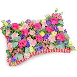 Seen from a gentle overhead angle on a pristine white backdrop, this classic rectangular floral pillow is densely packed with a lively mix of fresh blooms and greenery. Hot pink roses form the heart of the arrangement, their velvety petals producing a warm, affectionate focal point that is balanced by clusters of pale pink carnations for a soft, ruffled surface. Interspersed are purple lisianthus and sunlit lemon-yellow ranunculus that lift the composition with pops of contrasting colour, while small pink filler flowers add delicate punctuation. The distinctive silver-blue Eryngium thistles introduce a spiky, textural counterpoint, and generous stems of round, silvery eucalyptus leaves weave through the flowers, giving a cool scent and visual respite. A deep red, pleated satin ribbon trims the base in a ruffled border that defines the cushion silhouette and lends an elegant, memorial finish. The lighting is gentle and even, highlighting petal sheen and the subtle interplay of tones. Designed for moments of remembrance, whether placed at a funeral or in tribute at Golders Green Crematorium or a local church, this pillow reads as an expression of enduring affection and celebratory memory rather than stark grief. Crafted locally by Brent Cross Flowers, it reflects our neighbourhood care - ideal for families in Brent Cross, Cricklewood and nearby communities seeking a thoughtful, tasteful sympathy tribute.